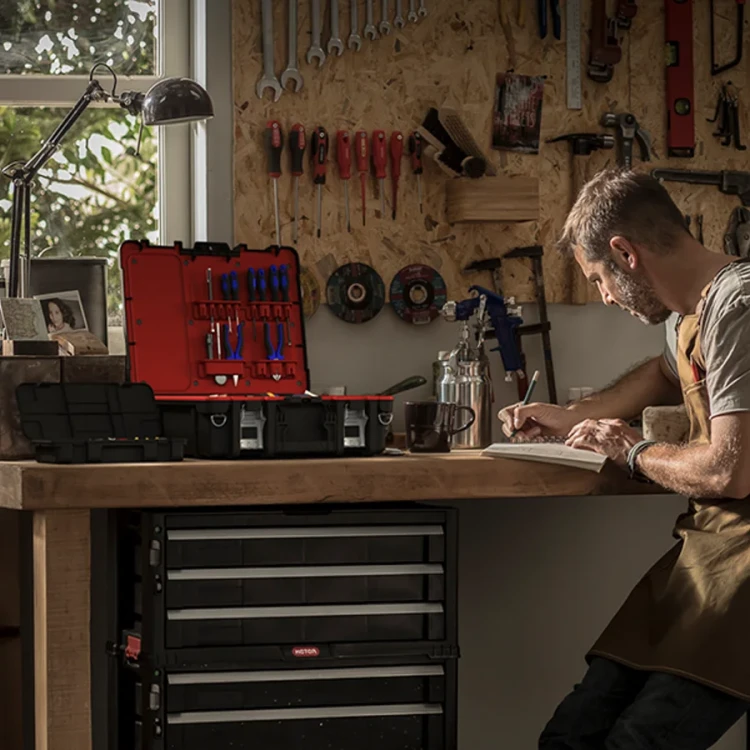Mężczyzna siedzący przy drewnianym blacie warsztatowym, otwarta walizka narzędziowa z czerwonym wnętrzem i narzędziami ustawiona na blacie obok dwóch zamkniętych pojemników, w tle ściana z zawieszonymi narzędziami.