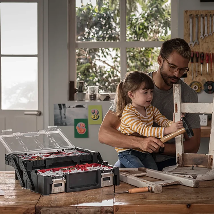 Mężczyzna i dziecko wspólnie naprawiają krzesło w domowym warsztacie; na stole obok otwarta, rozkładana czarno-czerwona skrzynka z przezroczystą pokrywą i czerwonymi przegródkami.