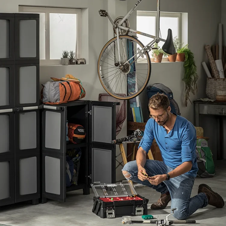 Mężczyzna klęczy w garażu i sortuje elementy w otwartej czarno-czerwonej skrzynce z przezroczystą pokrywą; w tle szafki, rower i akcesoria.