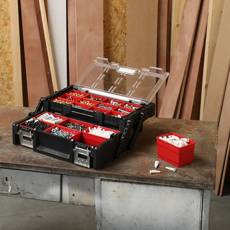 Otwarta skrzynka narzędziowa ustawiona na blacie warsztatowym, wewnątrz liczne czerwone przegródki z posortowanymi elementami; obok jeden z pojemników.