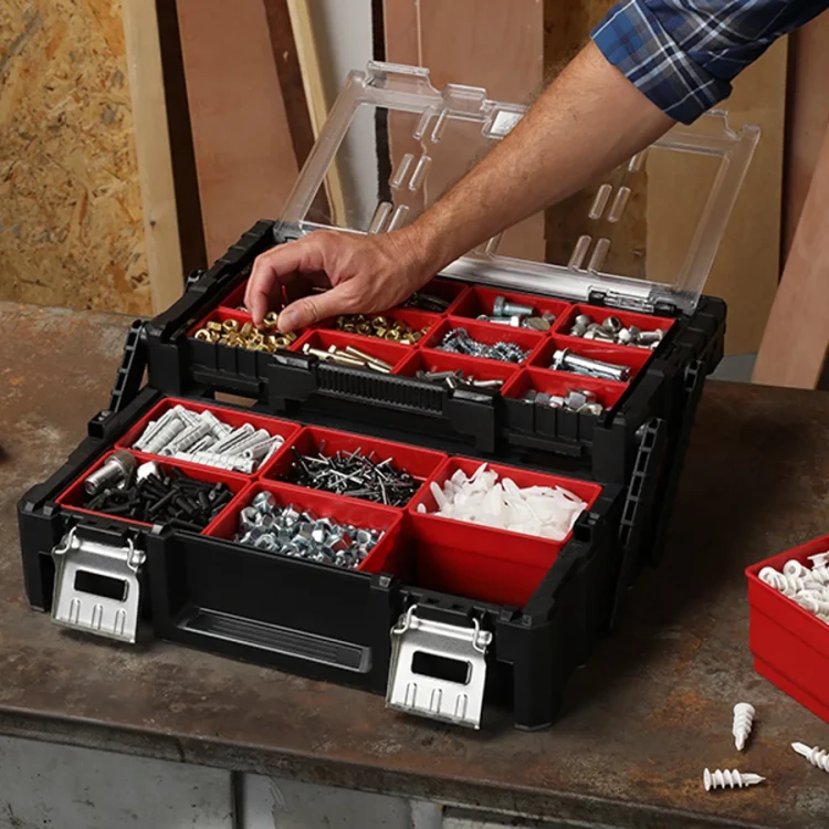 Widok z góry na otwartą skrzynkę narzędziową z przegródkami wypełnionymi różnymi elementami montażowymi; ręka sięga po śrubkę.