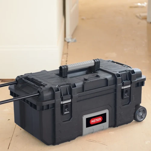 Skrzynka z rozłożonym uchwytem i kołami stoi na podłodze w pomieszczeniu, widok z przodu pod kątem.