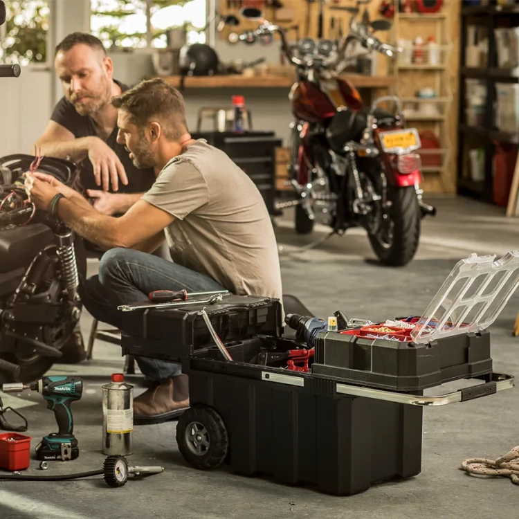 Dwóch mężczyzn pracuje przy motocyklu w przestronnym garażu; na pierwszym planie rozłożona czarna skrzynka narzędziowa z przezroczystą pokrywą, czerwonymi pojemnikami i metalową prowadnicą.