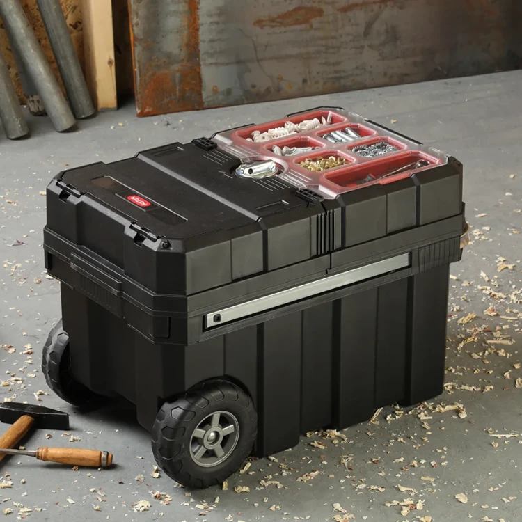 Skrzynka narzędziowa w zamkniętej pozycji, z widoczną przezroczystą pokrywą z przegrodami na drobne elementy, umieszczona na warsztatowej podłodze wśród wiórów.