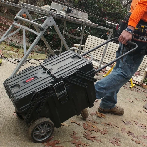 Osoba ciągnie skrzynkę po zewnętrznym podłożu za wysunięty metalowy uchwyt; w tle stół warsztatowy i narzędzia.
