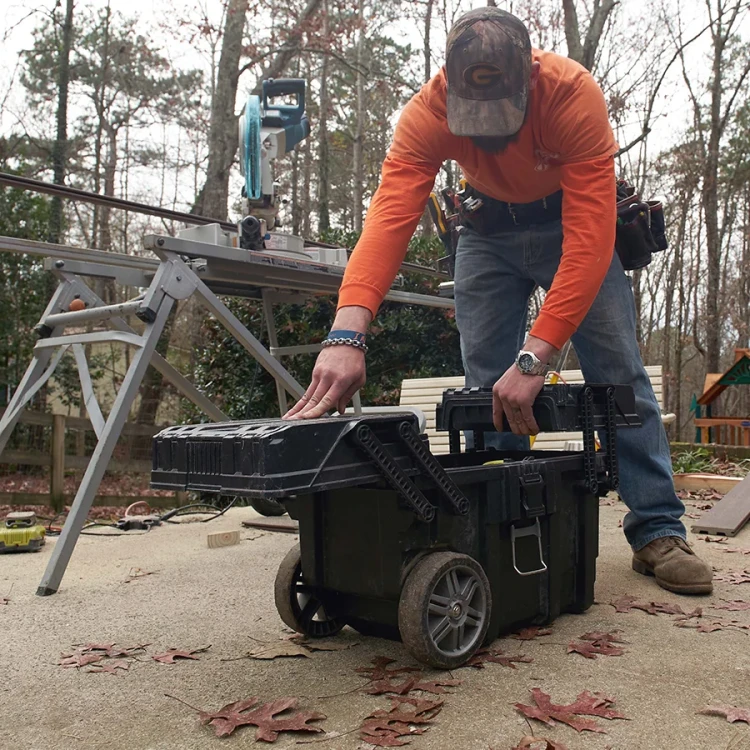 Mężczyzna w roboczym stroju otwiera skrzynkę narzędziową ustawioną na zewnątrz; pokrywa jest częściowo uniesiona, widoczne duże koła i mechanizm zawiasowy.