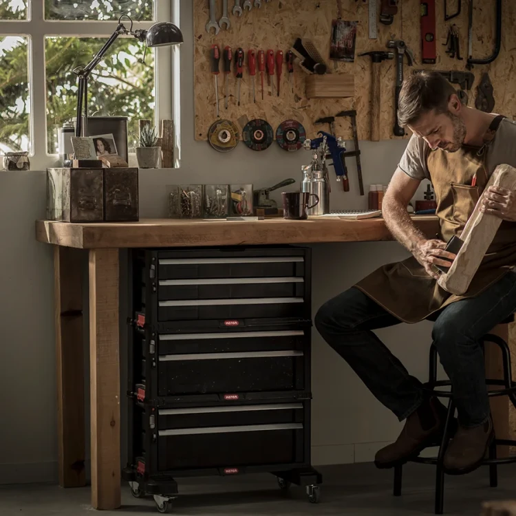 Regał z kilkoma modułami umieszczony pod drewnianym blatem roboczym w warsztacie, obok mężczyzna pracujący nad kawałkiem drewna.