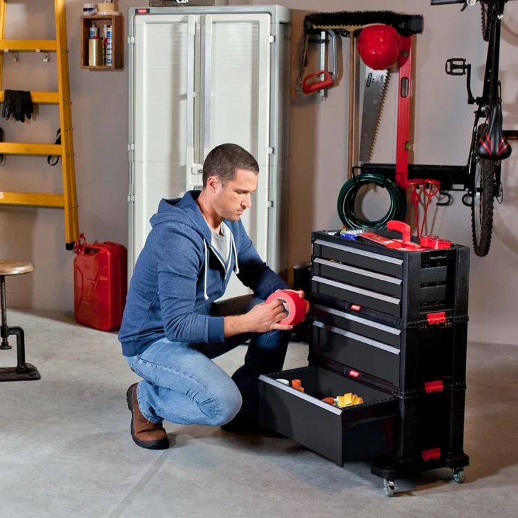 Mężczyzna w garażu przykuca przy regale z wysuniętą dolną szufladą, trzymając rolkę czerwonej taśmy.