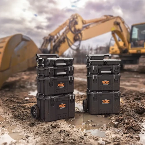 Dwie wieże złożone z czarnych skrzynek narzędziowych na tle placu budowy z koparką i błotnistym podłożem.